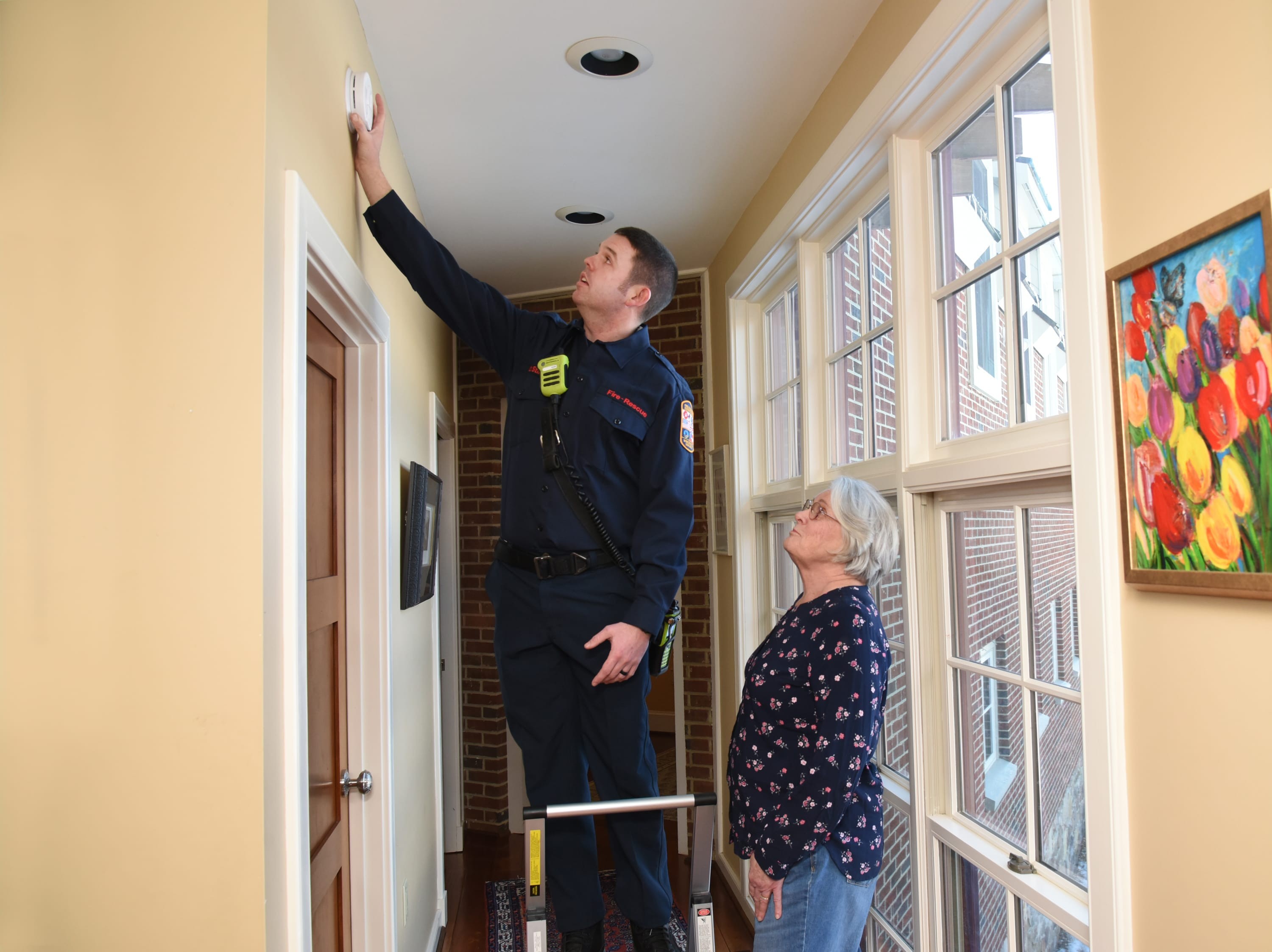 Firefighter testing a wall-mounted smoke alarm while a resident watches